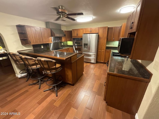 kitchen featuring tile countertops, light wood-style flooring, appliances with stainless steel finishes, a peninsula, and wall chimney range hood