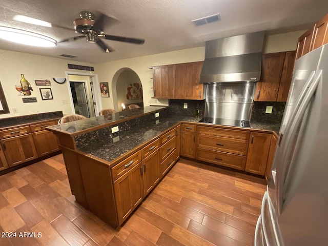 kitchen featuring tile counters, brown cabinetry, freestanding refrigerator, wall chimney range hood, and a peninsula