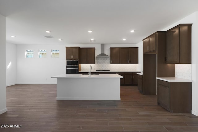 kitchen featuring dark wood-type flooring, stainless steel appliances, wall chimney range hood, sink, and a kitchen island with sink