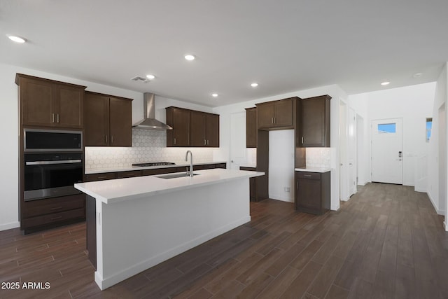 kitchen featuring a center island with sink, sink, wall chimney range hood, stainless steel appliances, and dark wood-type flooring