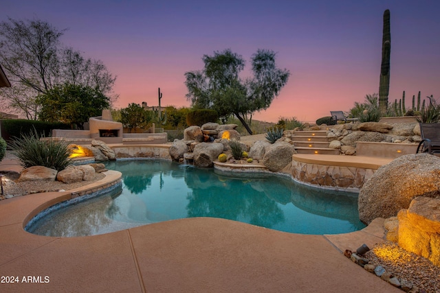 pool at dusk with pool water feature, an outdoor stone fireplace, and a patio