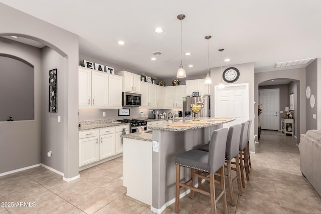 kitchen featuring hanging light fixtures, stainless steel appliances, light stone counters, an island with sink, and white cabinets