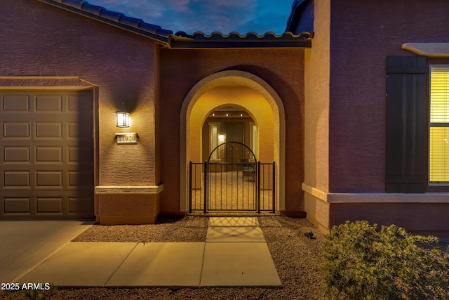 view of exterior entry with a tiled roof, an attached garage, a gate, and stucco siding