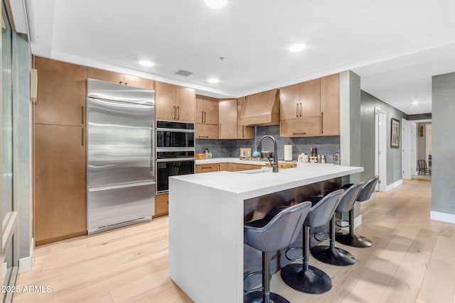 kitchen featuring light wood-type flooring, premium range hood, a breakfast bar, double oven, and built in refrigerator