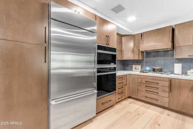 kitchen with custom exhaust hood, light hardwood / wood-style flooring, stainless steel appliances, and tasteful backsplash