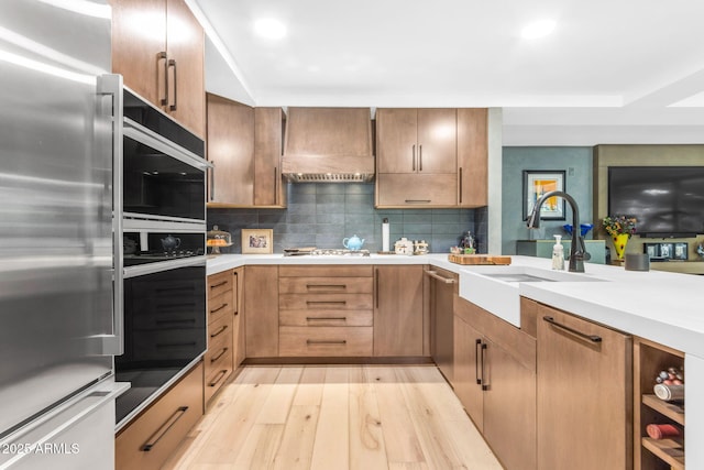 kitchen with sink, stainless steel appliances, decorative backsplash, custom range hood, and light wood-type flooring