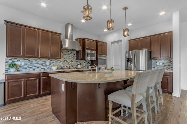 kitchen with a kitchen island with sink, wall chimney range hood, hanging light fixtures, sink, and appliances with stainless steel finishes