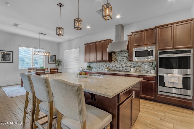 kitchen with a center island with sink, stainless steel appliances, light hardwood / wood-style floors, wall chimney exhaust hood, and decorative light fixtures