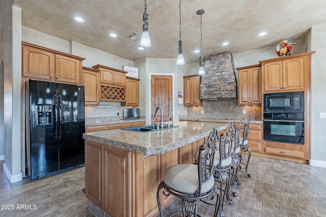 kitchen with visible vents, a kitchen island with sink, a sink, wall chimney range hood, and black appliances