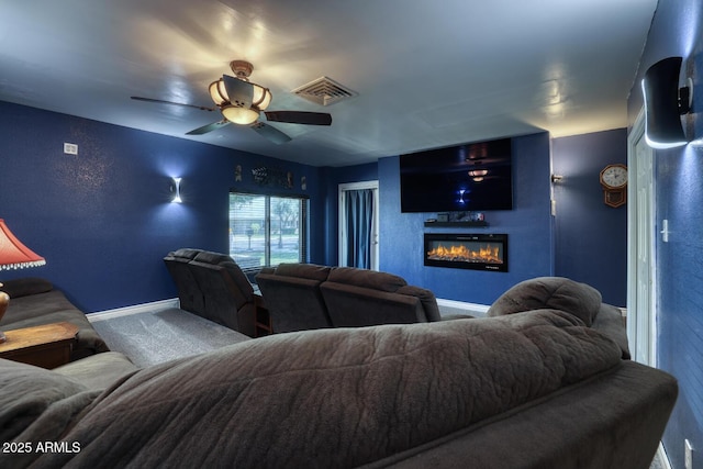 carpeted home theater room featuring visible vents, baseboards, a glass covered fireplace, and ceiling fan