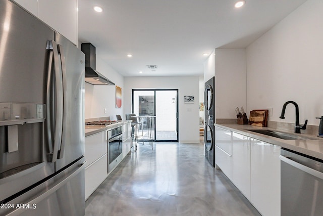 kitchen with sink, white cabinets, wall chimney range hood, stainless steel appliances, and concrete flooring