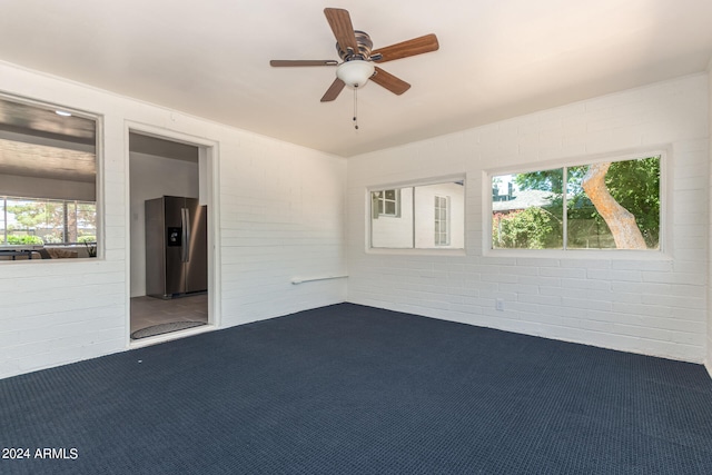 carpeted empty room with a healthy amount of sunlight, ceiling fan, and brick wall