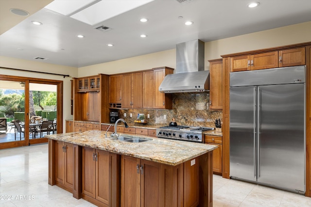 kitchen with light stone countertops, high end appliances, sink, wall chimney range hood, and a center island with sink