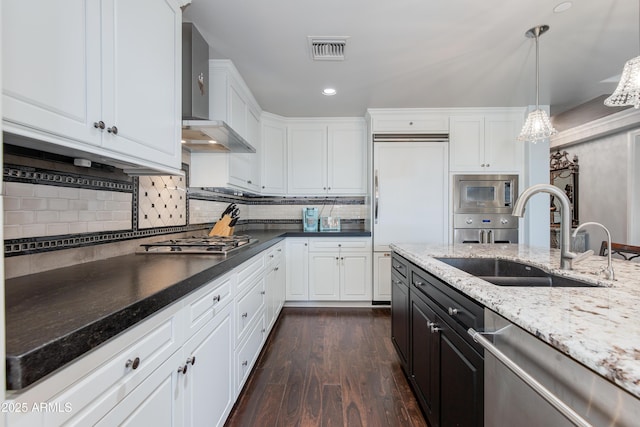 kitchen featuring visible vents, a sink, white cabinets, built in appliances, and wall chimney exhaust hood