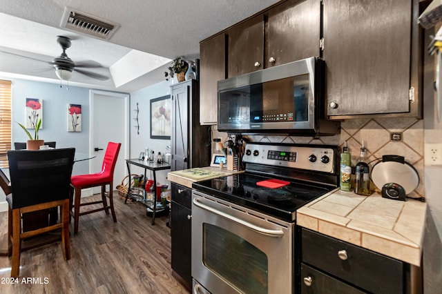 kitchen with dark brown cabinets, stainless steel appliances, ceiling fan, dark hardwood / wood-style floors, and tile counters