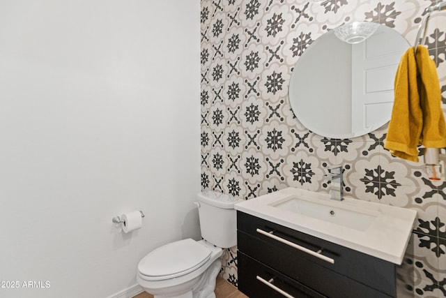 half bathroom with vanity, baseboards, toilet, and wallpapered walls