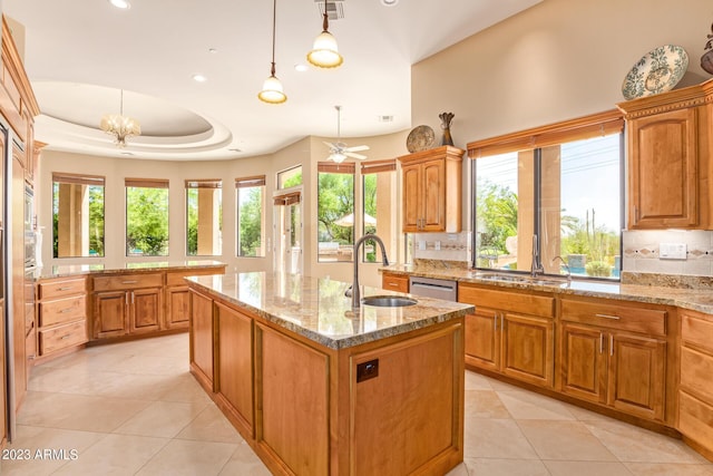 kitchen with light stone countertops, hanging light fixtures, sink, a raised ceiling, and a center island with sink