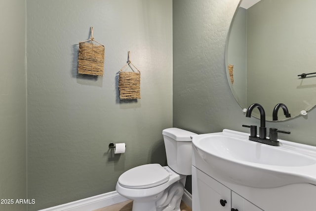 half bath featuring toilet, a textured wall, baseboards, and vanity