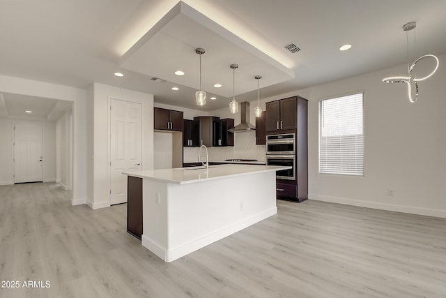 kitchen with decorative light fixtures, light wood-type flooring, black electric stovetop, a kitchen island with sink, and wall chimney range hood