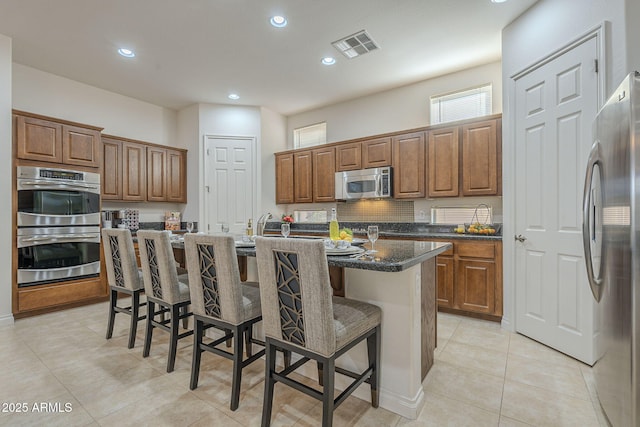 kitchen with a breakfast bar area, a center island with sink, light tile patterned floors, appliances with stainless steel finishes, and dark stone counters