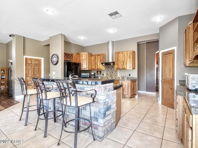 kitchen with wall chimney exhaust hood, black refrigerator with ice dispenser, a breakfast bar, light tile patterned floors, and a center island with sink