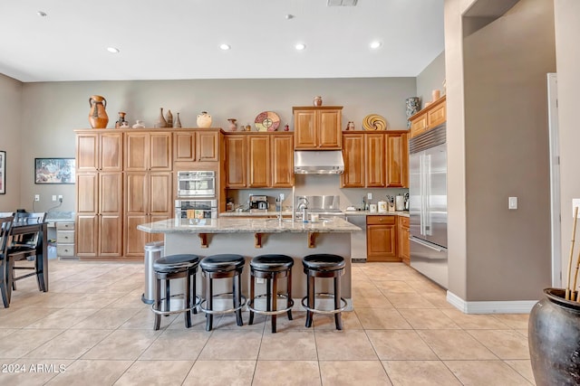 kitchen featuring light tile patterned floors, light stone counters, under cabinet range hood, stainless steel appliances, and an island with sink