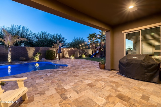 pool at dusk featuring a patio area, grilling area, a playground, and a fenced backyard