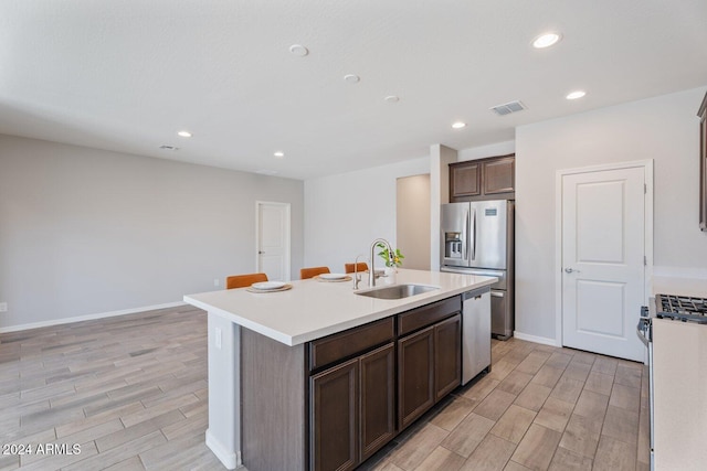 kitchen with dark brown cabinetry, sink, stainless steel appliances, light hardwood / wood-style floors, and a center island with sink