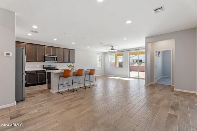 kitchen with a kitchen bar, appliances with stainless steel finishes, light wood-type flooring, a kitchen island with sink, and ceiling fan