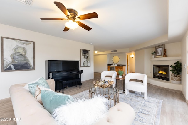 living room with a glass covered fireplace, visible vents, light wood finished floors, and ceiling fan