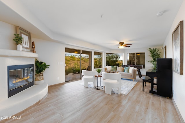 living room featuring light wood-type flooring, baseboards, a glass covered fireplace, and a ceiling fan