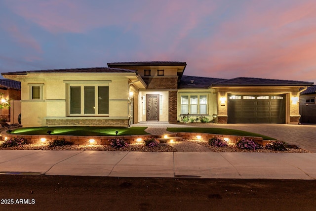 prairie-style house with a garage, brick siding, decorative driveway, and stucco siding