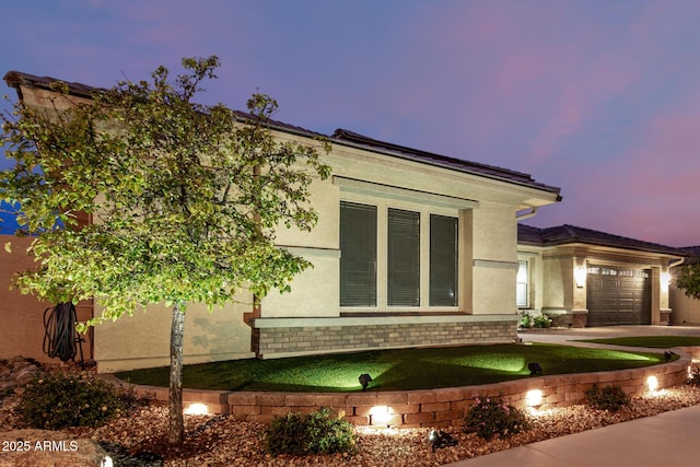 property exterior at dusk with an attached garage, brick siding, driveway, and stucco siding