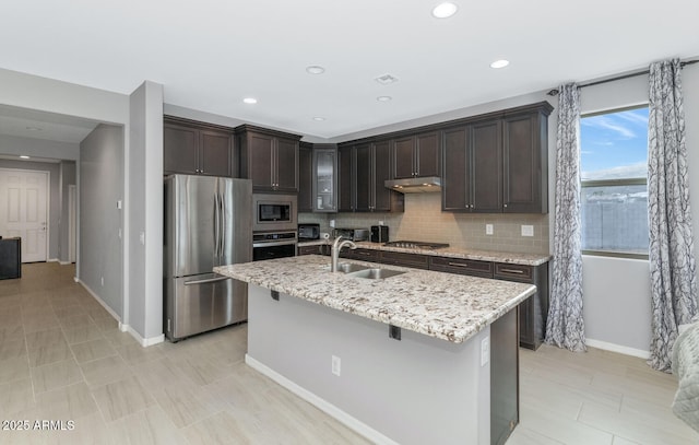 kitchen featuring sink, light stone countertops, an island with sink, appliances with stainless steel finishes, and dark brown cabinets