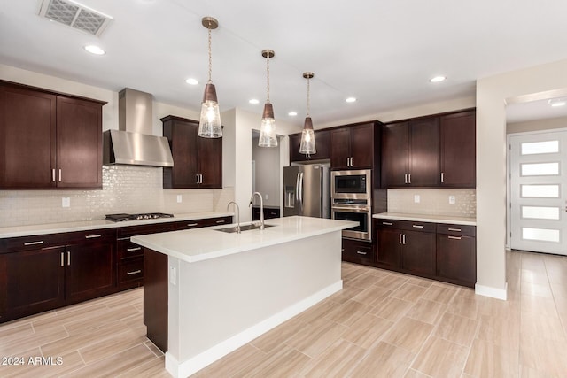 kitchen featuring wall chimney exhaust hood, stainless steel appliances, a kitchen island with sink, sink, and pendant lighting