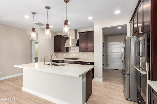 kitchen with wall chimney range hood, sink, light hardwood / wood-style flooring, an island with sink, and stainless steel appliances