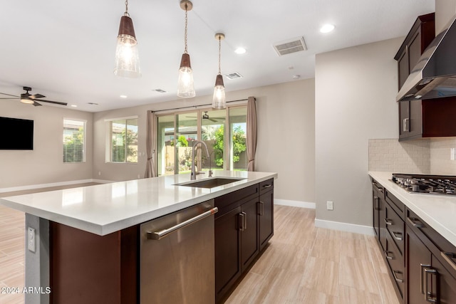 kitchen with ceiling fan, sink, wall chimney exhaust hood, a kitchen island with sink, and appliances with stainless steel finishes