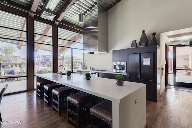 kitchen featuring dark wood-style flooring, a towering ceiling, light countertops, appliances with stainless steel finishes, and an island with sink