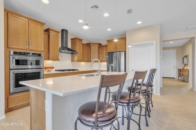 kitchen featuring sink, an island with sink, pendant lighting, stainless steel appliances, and wall chimney range hood