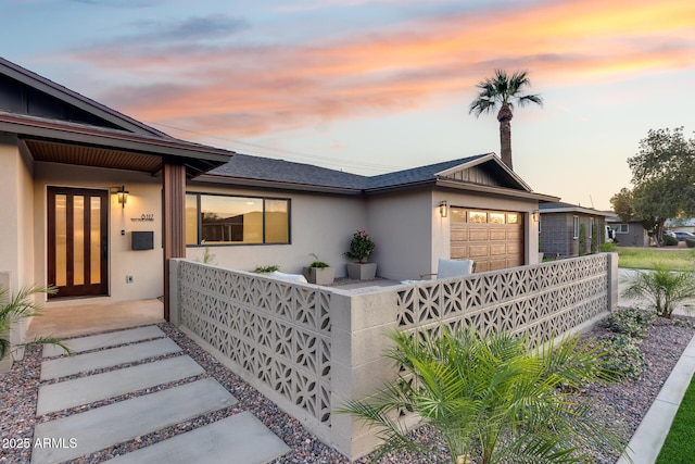 view of front of house featuring an attached garage, roof with shingles, stucco siding, and fence