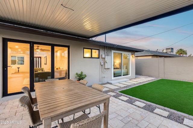 back of property at dusk featuring fence, outdoor dining space, stucco siding, a patio, and a yard