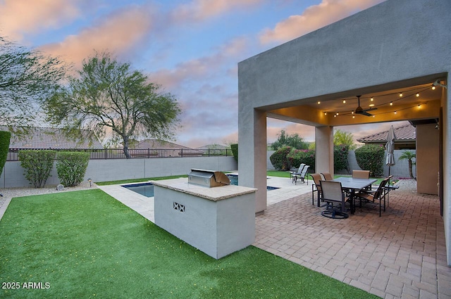 patio terrace at dusk featuring ceiling fan, a lawn, exterior kitchen, a fenced backyard, and outdoor dining space
