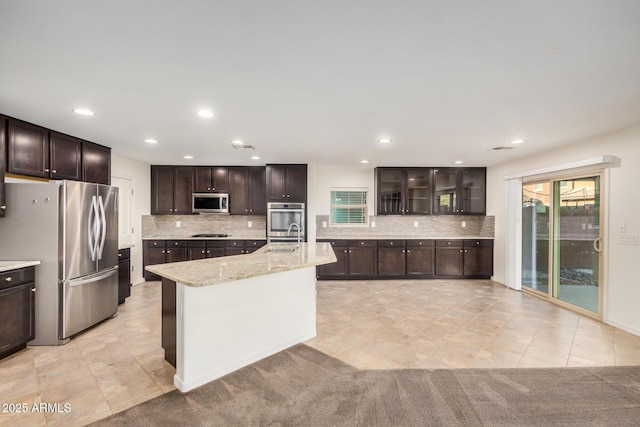 kitchen featuring decorative backsplash, glass insert cabinets, appliances with stainless steel finishes, a kitchen island with sink, and dark brown cabinets