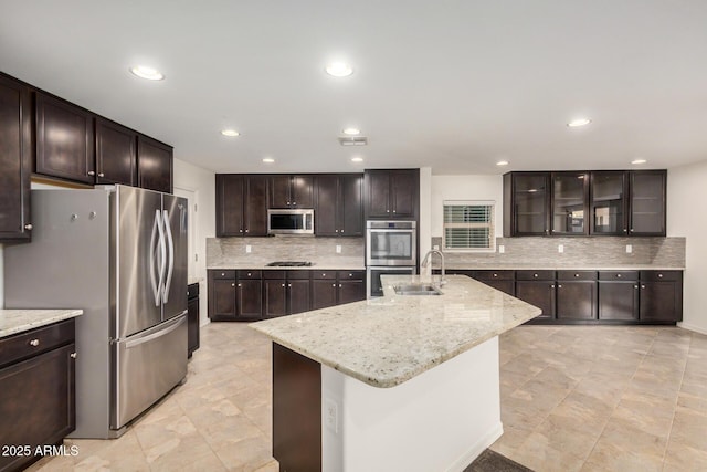 kitchen with dark brown cabinetry, stainless steel appliances, a sink, light stone countertops, and a center island with sink