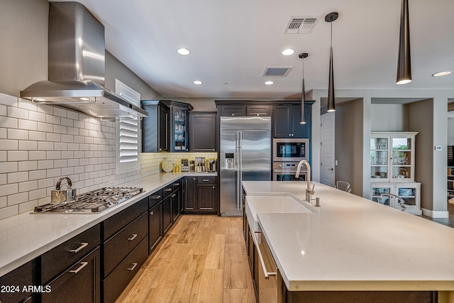 kitchen with decorative light fixtures, wall chimney range hood, built in appliances, backsplash, and a kitchen island with sink