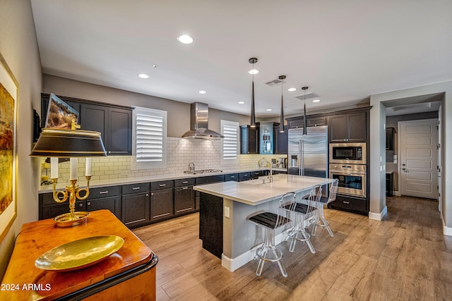 kitchen featuring wall chimney exhaust hood, built in appliances, hanging light fixtures, a breakfast bar, and a center island with sink