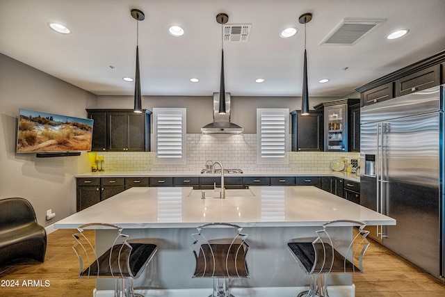 kitchen with sink, a center island with sink, high end fridge, and wall chimney range hood