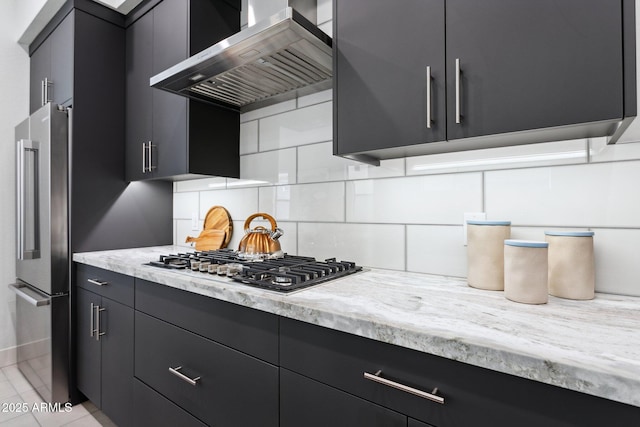 kitchen featuring dark cabinetry, wall chimney exhaust hood, stainless steel appliances, baseboards, and decorative backsplash