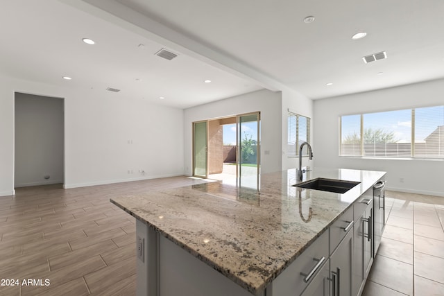 kitchen featuring sink, light stone countertops, an island with sink, stainless steel dishwasher, and gray cabinets