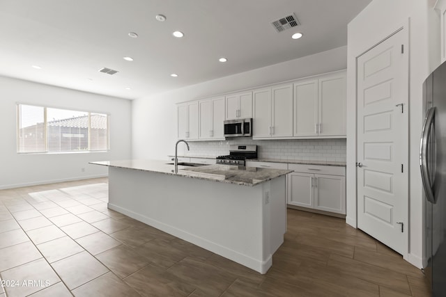 kitchen featuring white cabinetry, appliances with stainless steel finishes, light stone countertops, a kitchen island with sink, and decorative backsplash